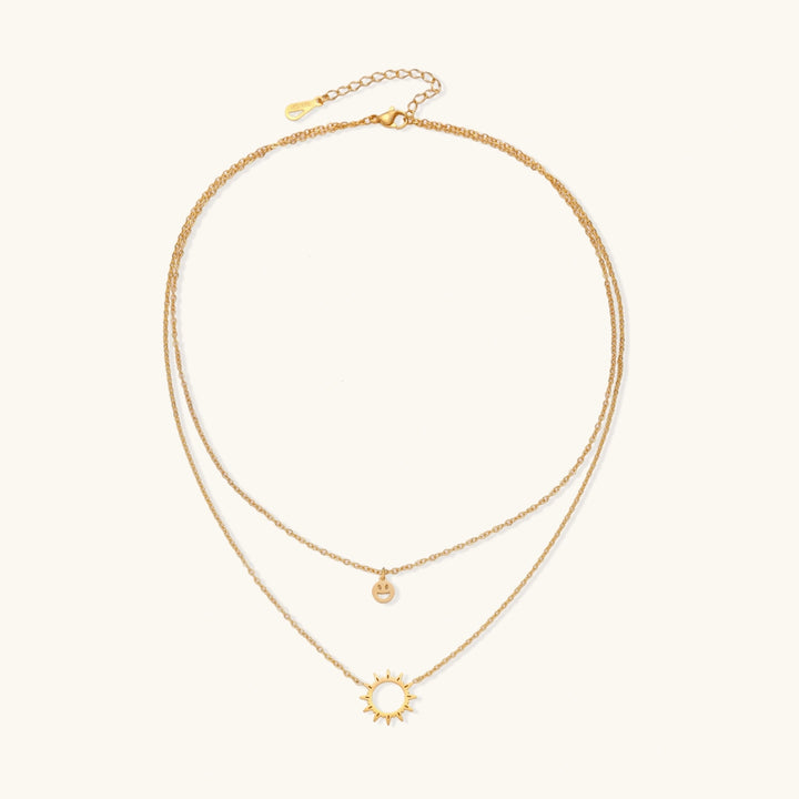 Gold layered necklace set with sunburst and smiley face pendants on delicate chains against white background.