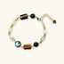 Gold chain bracelet with tiger's eye, obsidian, pearl, and iridescent abalone stones on cream background.