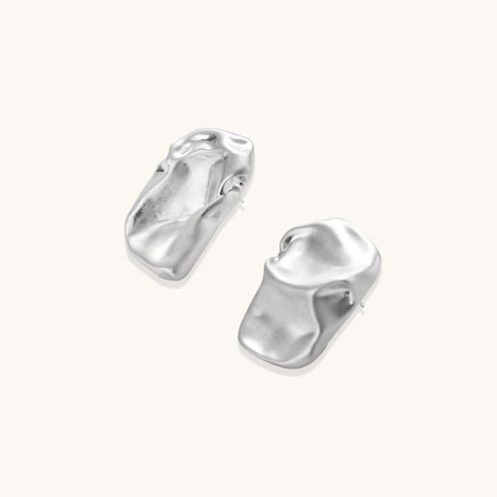 Pair of sculptural silver ear cuffs with organic fluid shapes and polished finish against cream background.