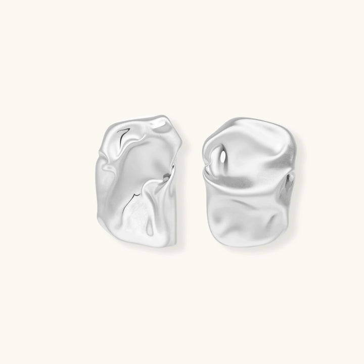 Pair of sculptural silver ear cuffs with organic fluid shapes and polished finish against cream background.