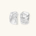 Pair of sculptural silver ear cuffs with organic fluid shapes and polished finish against cream background.